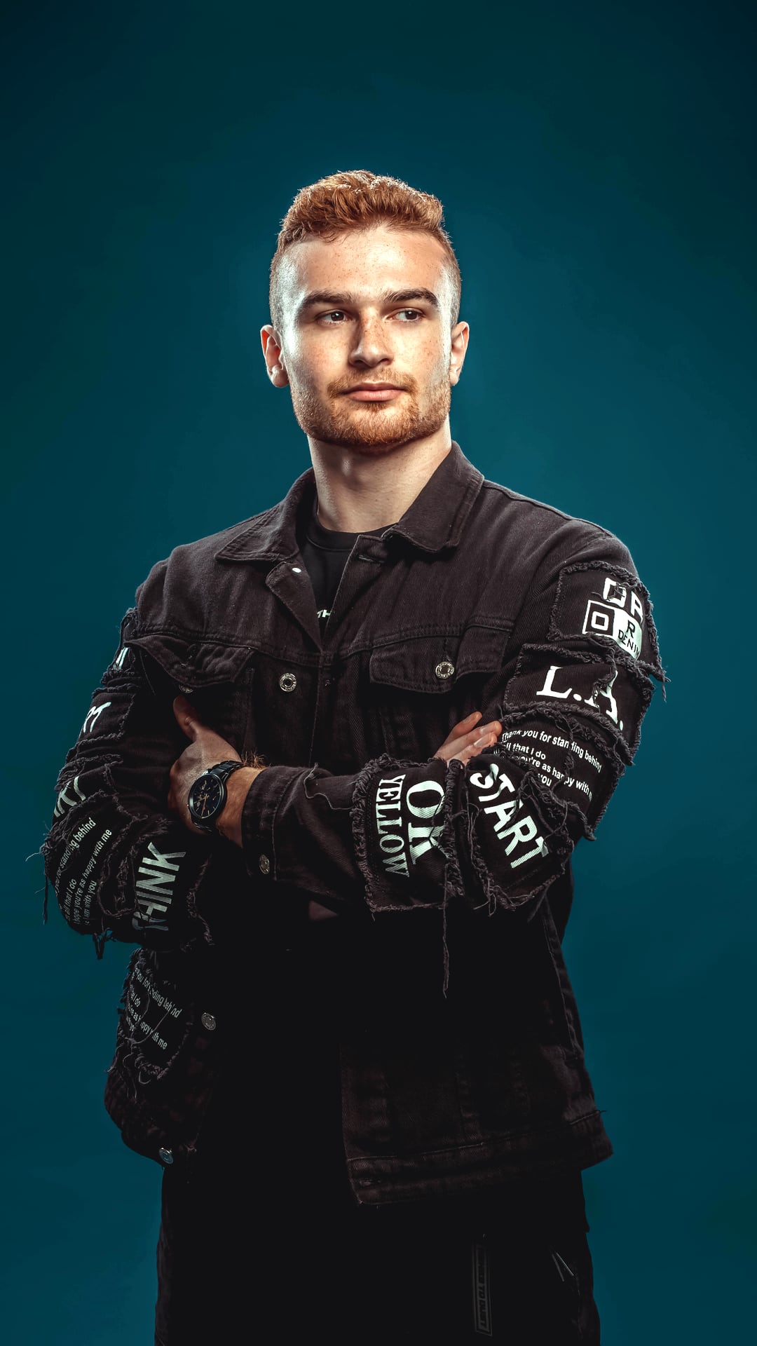 A-RIZE — Carlos van Boekhold, arms folded, in a black patched denim jacket against a petrol studio backdrop